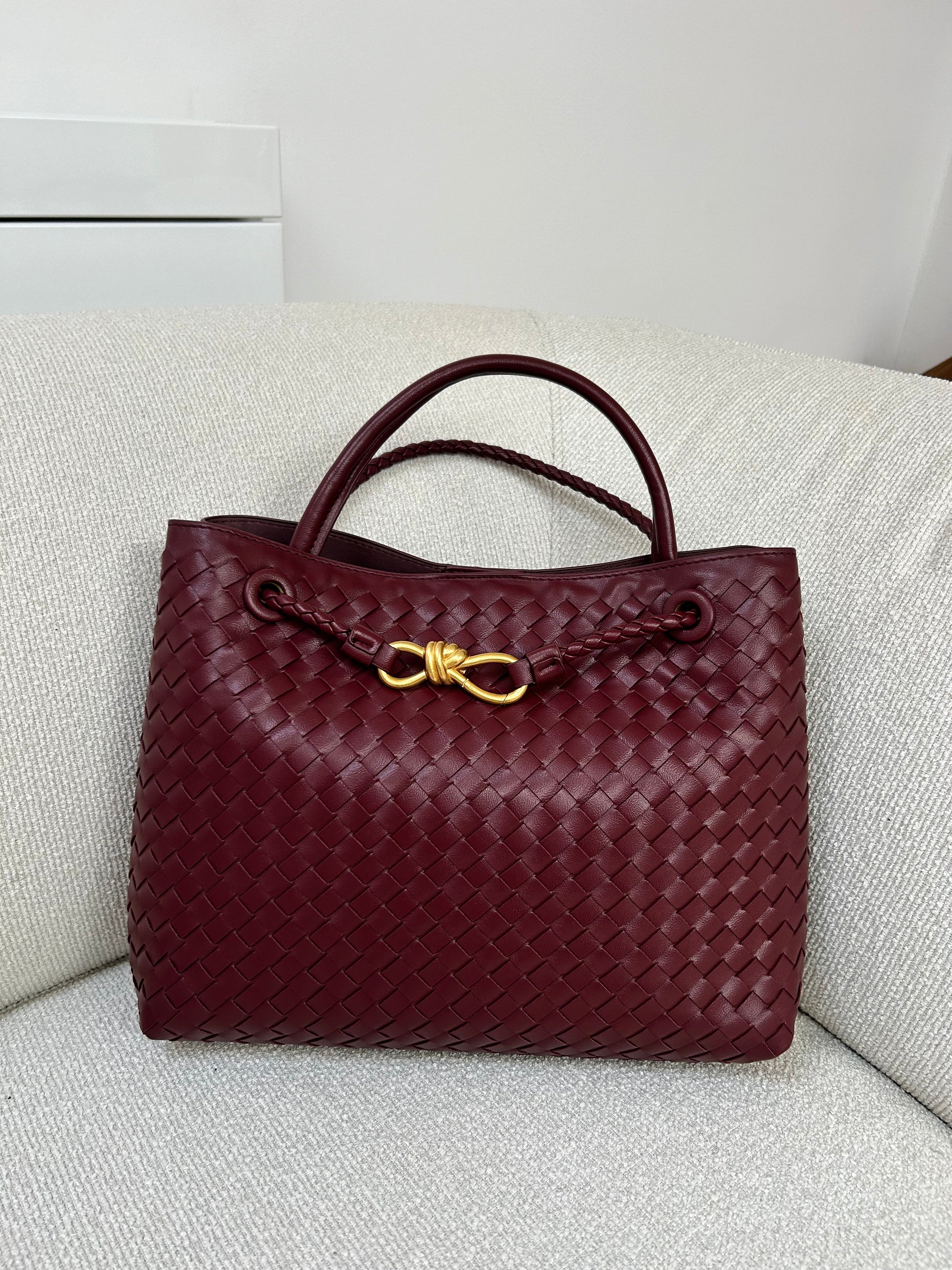 A burgundy elegant hand-woven bag with intricate braided patterns is placed on a stylish beige sofa.The rich deep red tone of the bag contrasts beautifully with the soft,neutral upholstery,creating a sophisticated and inviting scene.designer dupe bag