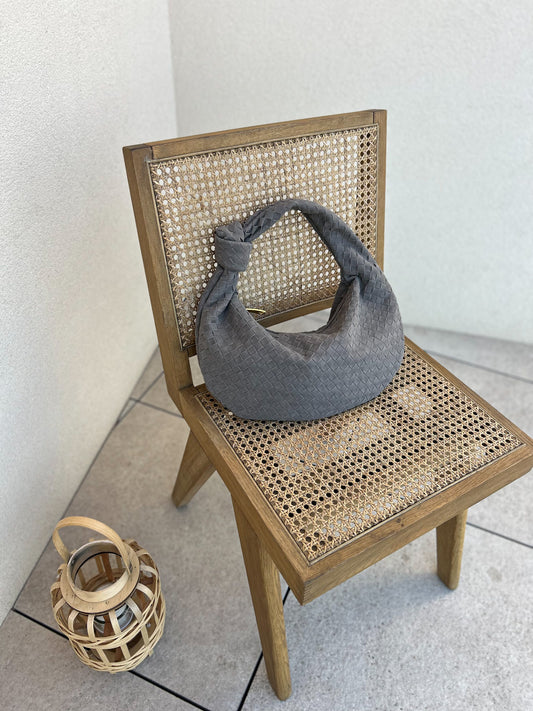 Grey vegan suede shoulder bag resting on a straw chair, showcasing its sleek and soft texture. The minimalist design of the bag contrasts beautifully with the natural texture of the chair, creating a chic, relaxed aesthetic for everyday style. pattyb