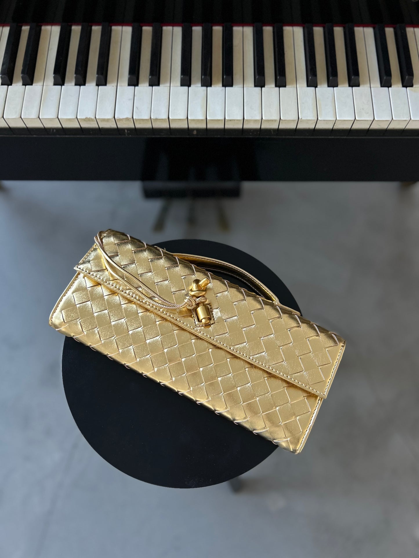 The Dumpling Purse design is a best seller loved for its unique style and high-quality craftsmanship, making it the perfect gift for the fashionable woman in your life. Upgrade your look with this must-have statement piece or treat your loved one - birthday, Mother's Day or as an everyday gift.
Gold woven clutch elegantly placed on a stool next to a piano.