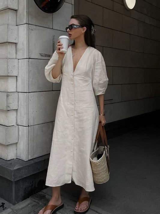 Woman wearing a cream linen midi dress with an open V-back and tie detail, styled with puff sleeves and flat leather sandals. She holds a coffee cup while walking through a modern city street, showcasing the dress’s relaxed and feminine silhouette—perfect for effortless summer styling.